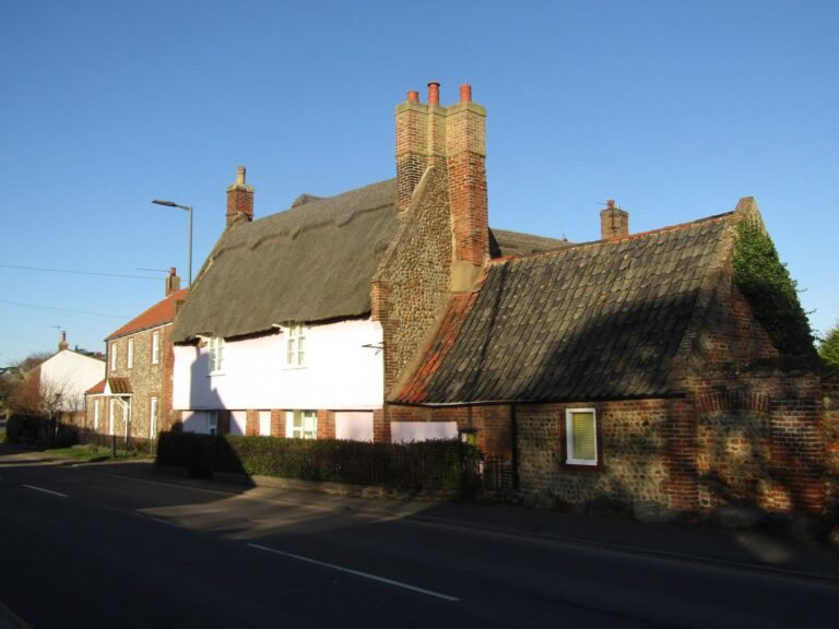 Thatched cottage with pale pink rendered walls beside flint and brick buildings on Walcott Road, Bacton